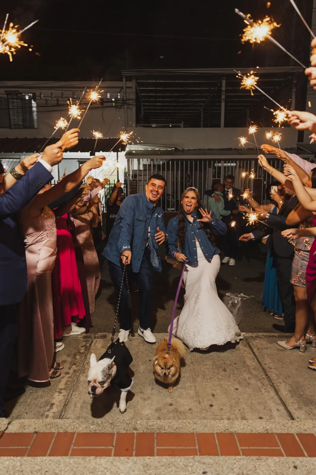 Pareja con chaquetas de denim caminando entre invitados con luces de bengala